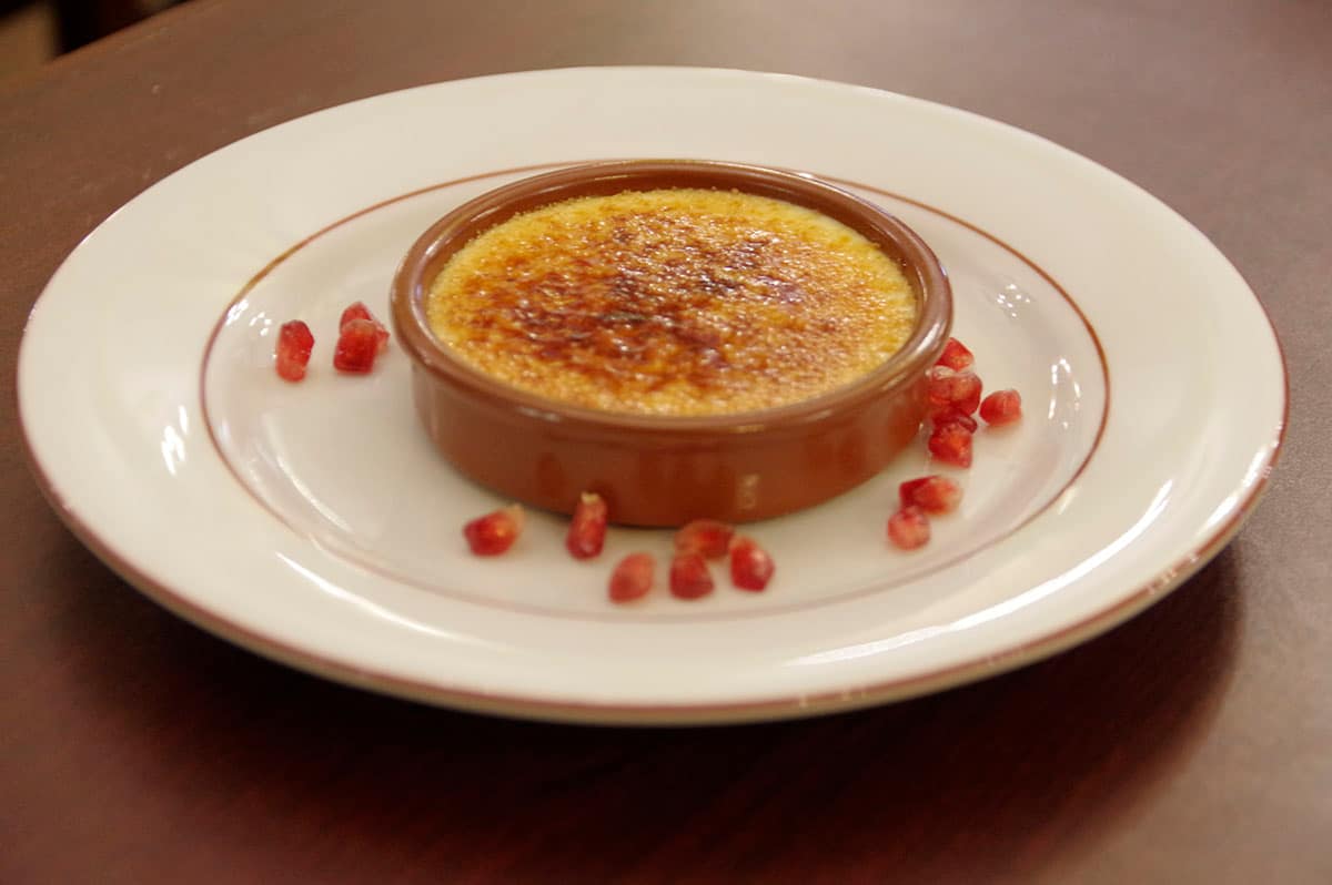 Crème brûlée caramélisée dans son plat en terre, parsemée de grains de grenade.