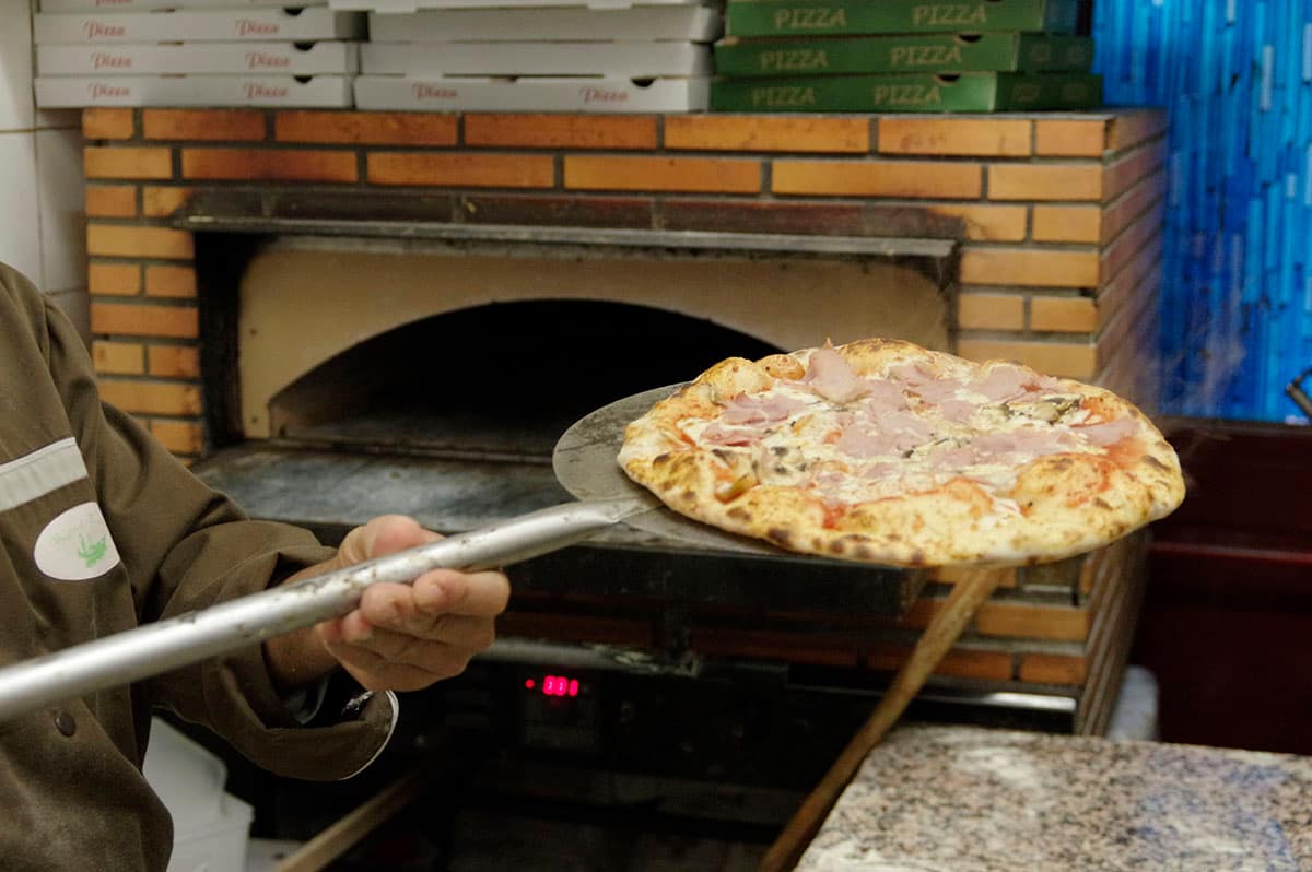 Une pizza sortie du four à l’aide de la pelle, devant le four à pizza du restaurant.