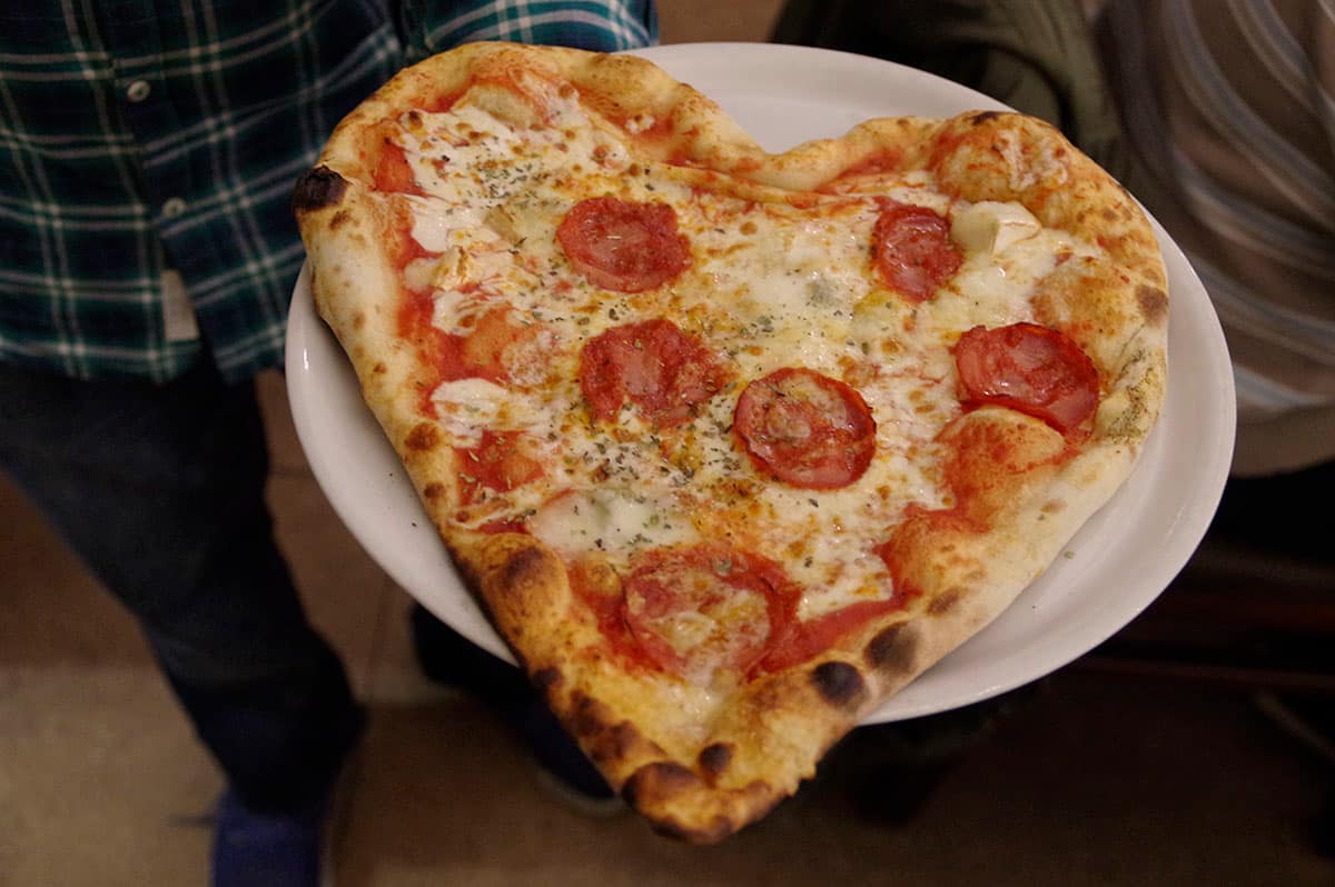 Pizza en forme de cœur, mozzarella et tomates, portée à table sur son assiette.
