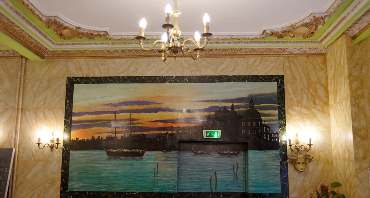 Peinture murale d’un coucher de soleil sur Venise, encadrée par la corniche ancienne et le marbre en trompe-l’œil de la salle.