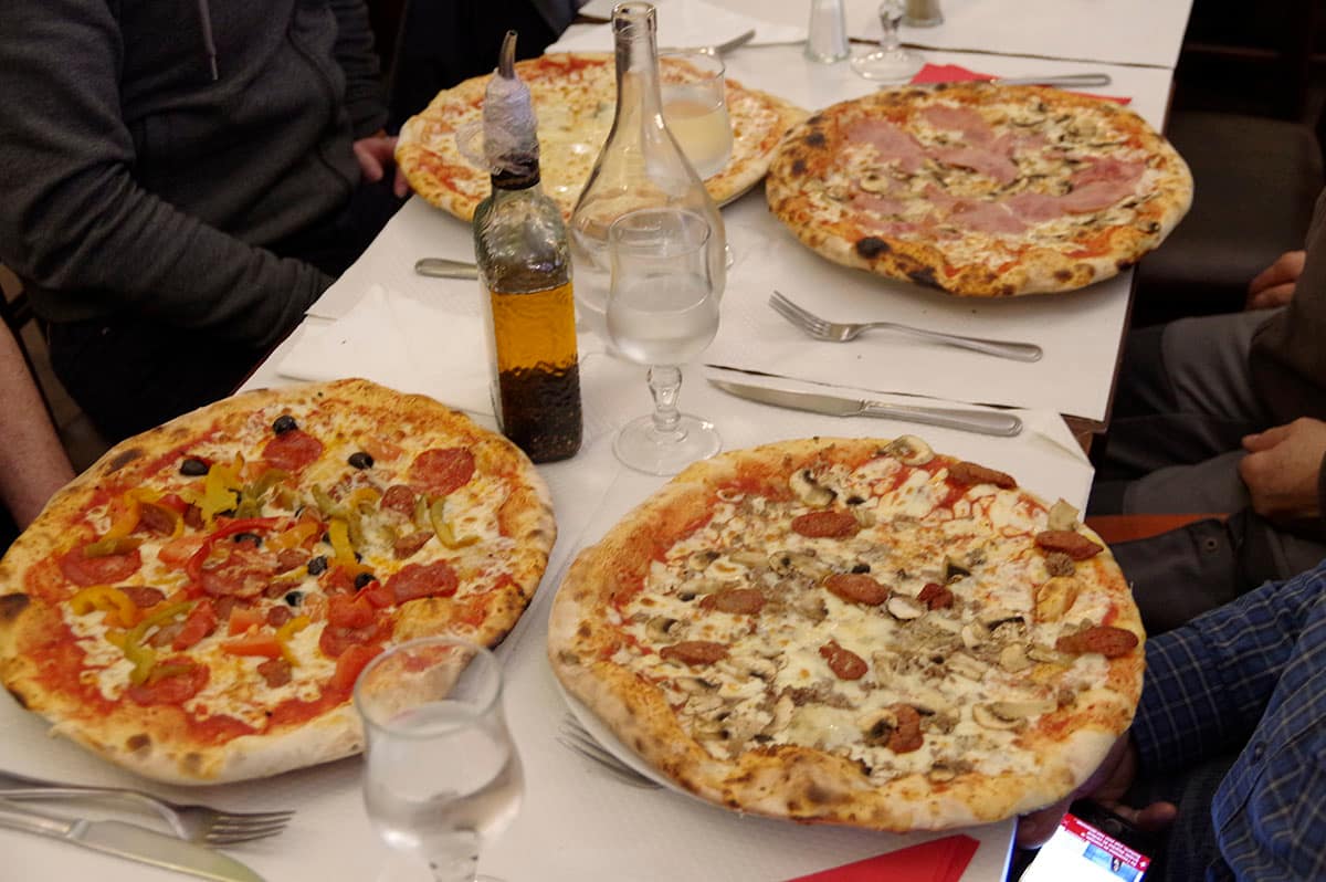 Quatre pizzas posées sur une grande table, entre les verres et la bouteille d’huile d’olive.